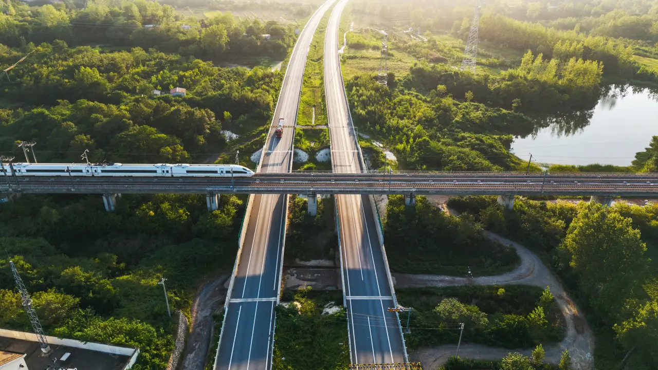 A high-speed train crossing a viaduct over a dual carriageway, with power lines and a lake